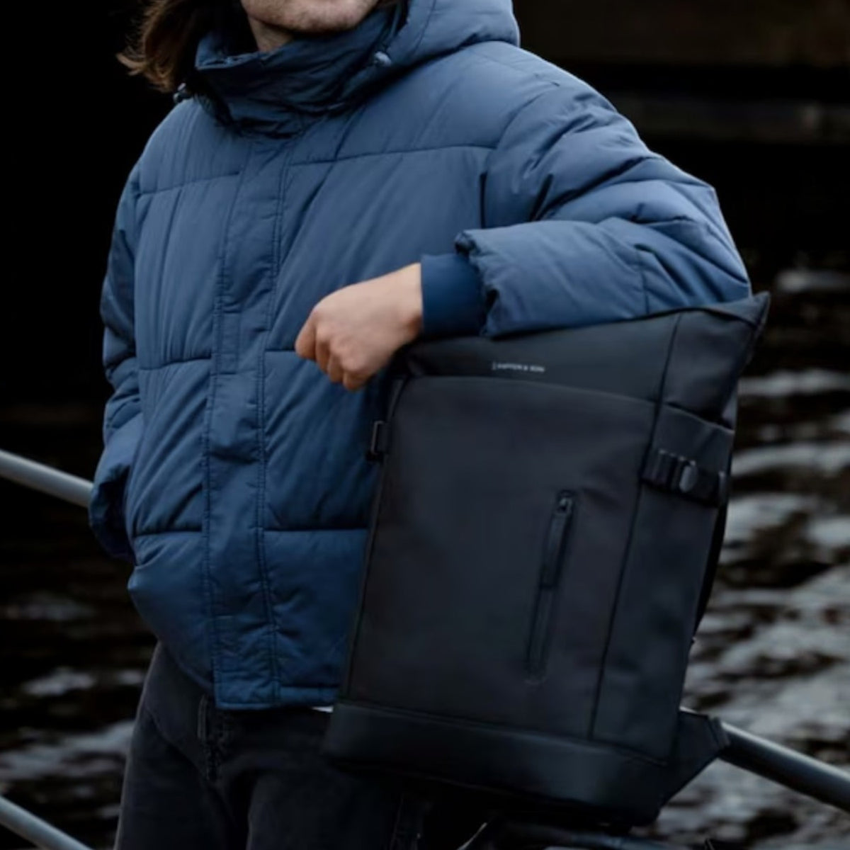 Man wearing a blue puffer jacket and black beanie posing with a Lieblingstasche Helsinki black minimalist backpack