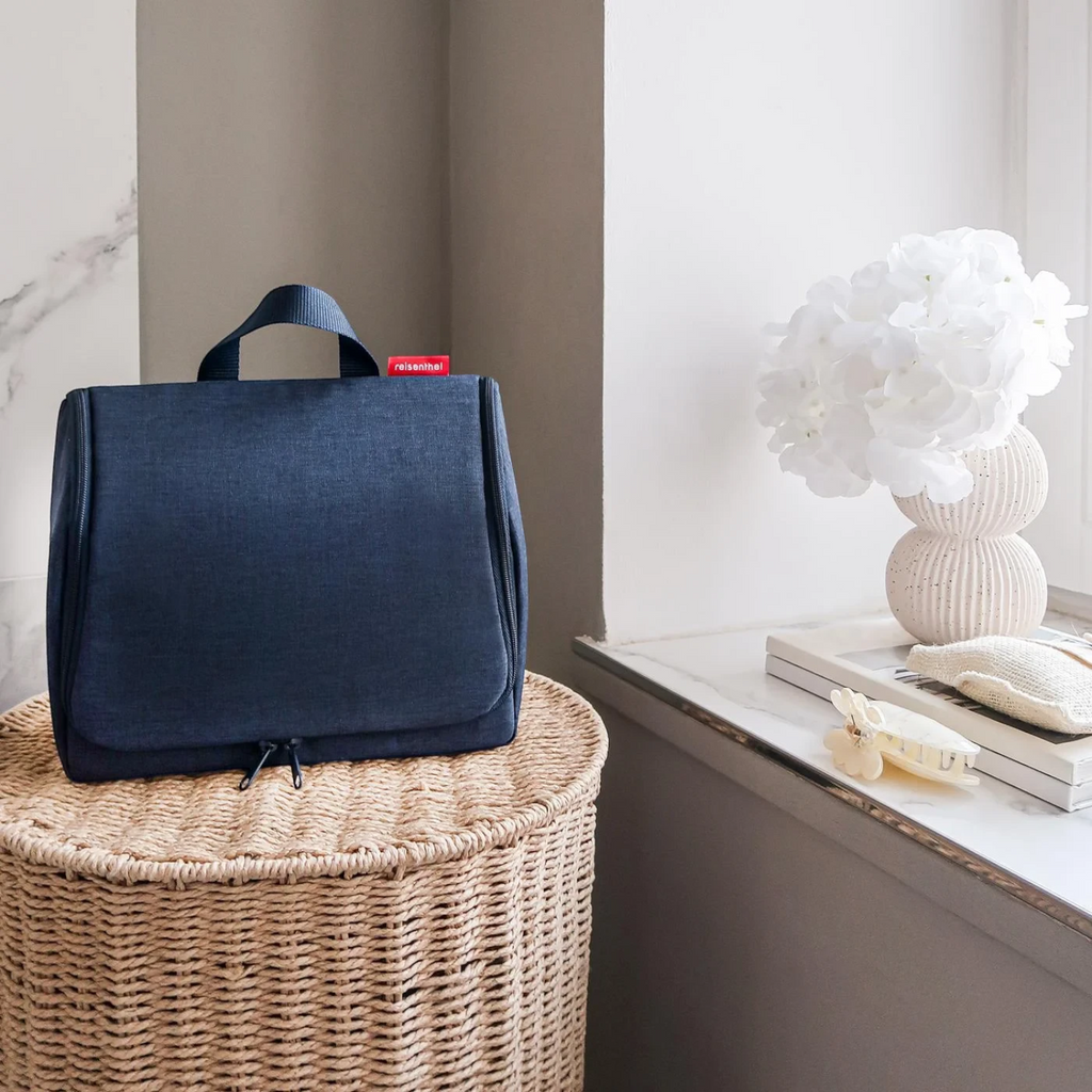 Lieblingstasche Toiletbag XL in dark blue fabric with a top handle, sitting on a woven basket near a white vase.